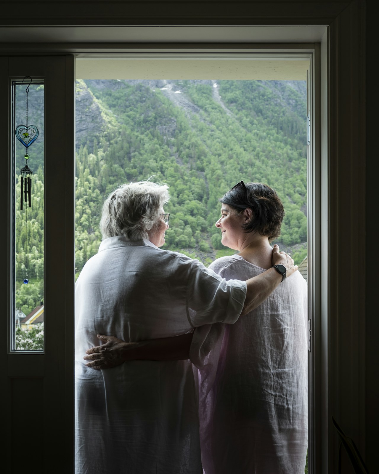 To kvinner, Undis og Maryan, kledd i hvitt, holder rundt hverandre mens de står i en åpne verandadør og ser ut på grønn natur.