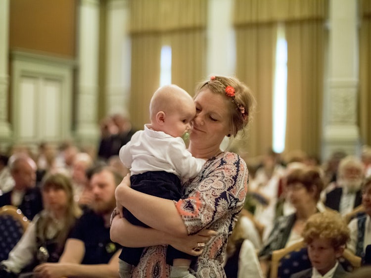 En mor holder sin baby tett inntil seg i en sal fylt med mennesker.
