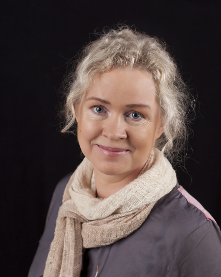 Portrettfoto av Ingun Steen Andersen. Kvinne med lys hud og lys blondt hår smiler. Grå topp, beige skjerf, svart bakgrunn.